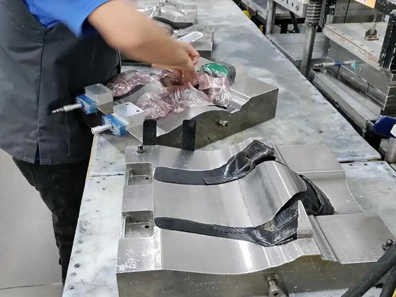 Image shows HANS device in production. Location is a manufacturing area with a row of large metal tools, with the shape of the HANS device embedded, sitting on a table top. The tool in the foreground has plys of prepreg laid up in the tool. Behind this, a technician is working on another tool, preparing the mold for processing.